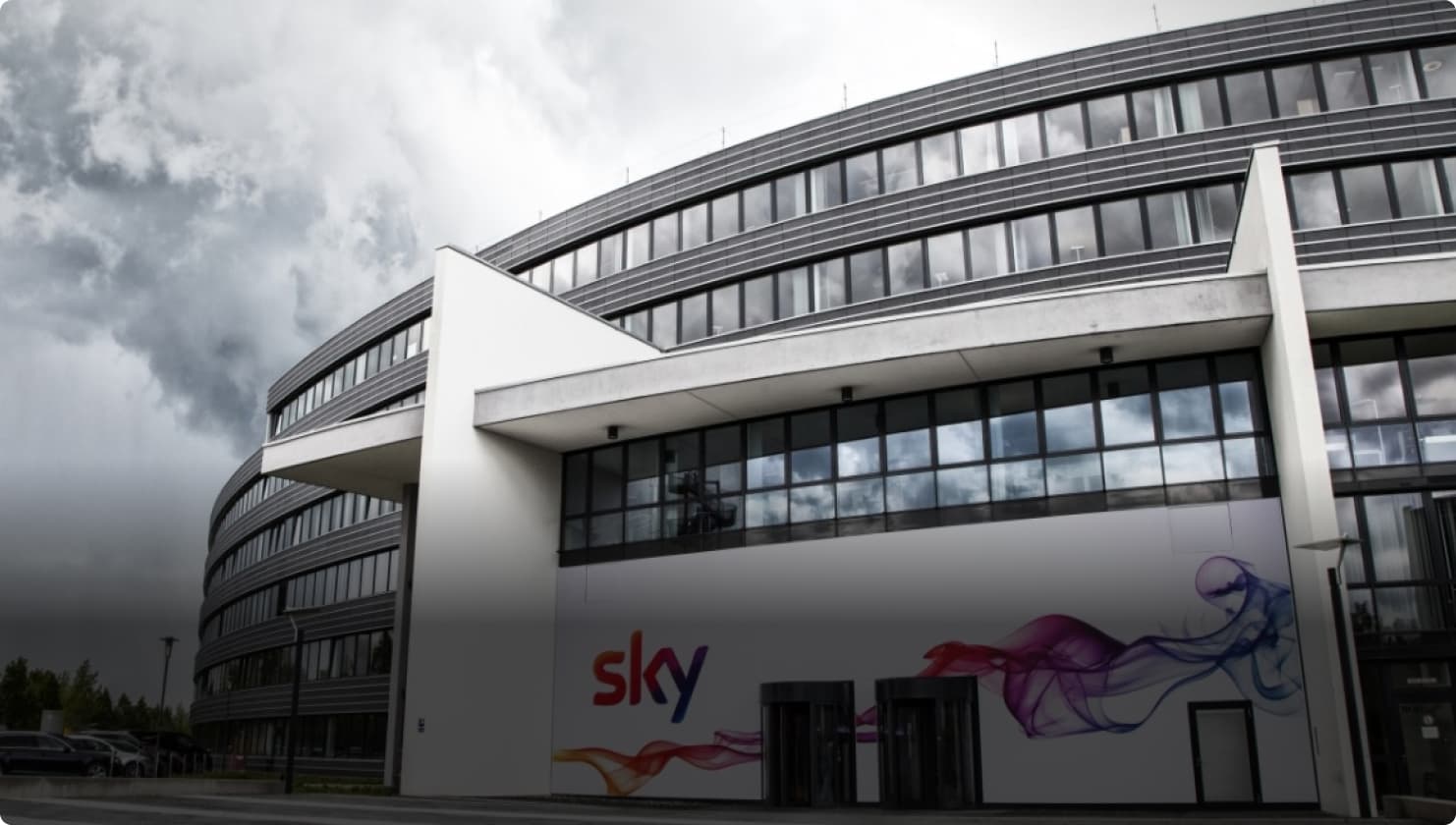 Exterior of a modern Sky office building with glass windows and a Sky logo wall mural beneath a cloudy sky.