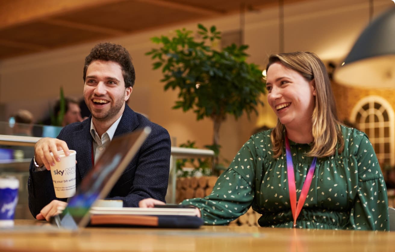Man and woman laughing during a conversation at a wooden table in an open office café area.