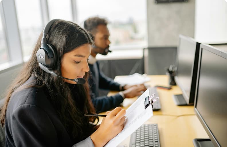 Woman in a call centre headset writing notes on a clipboard at her desk, with a male colleague beside her working on a desktop computer.