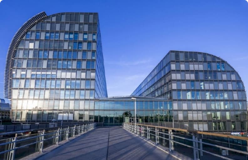 Modern glass office building with curved facades and a central walkway leading to a main entrance, set against a clear blue sky.