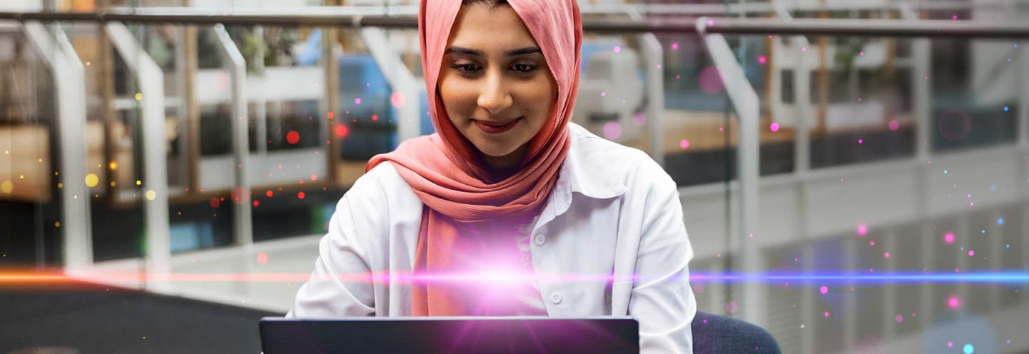 A woman in a pink hijab works on a laptop, surrounded by colorful glowing effects.
