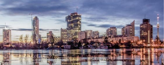 City skyline at dusk, buildings reflecting on calm water under a cloudy sky.