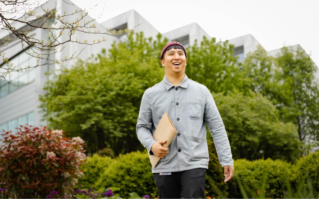 Man smiling and walking through a garden area outside a Sky building, holding documents.
