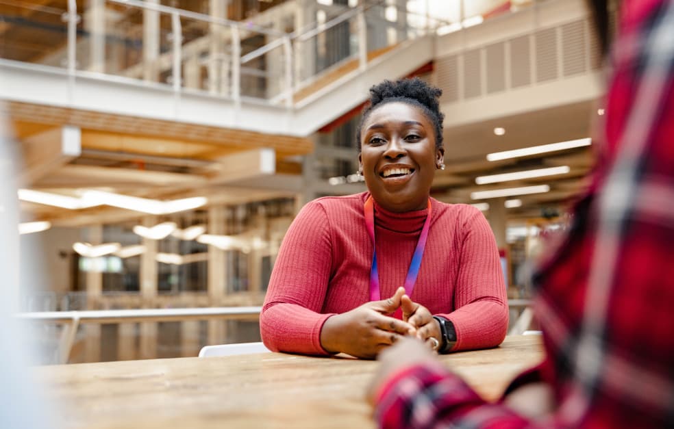 Woman smiling and sitting at a wooden table in a spacious indoor office atrium, speaking with someone across from her.