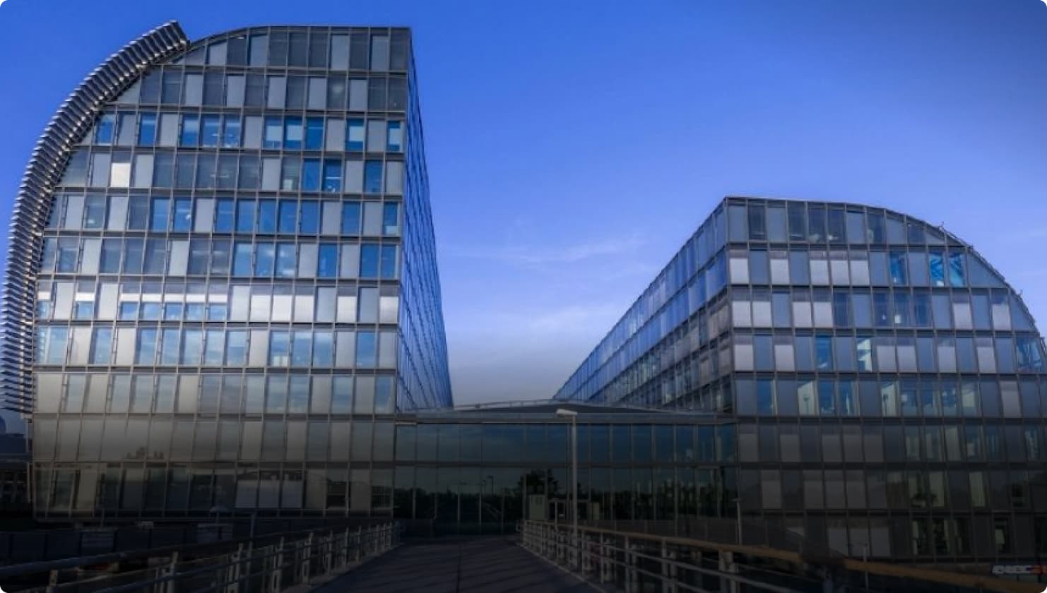 Modern glass office building with curved facades and a central walkway leading to a main entrance, set against a clear blue sky.