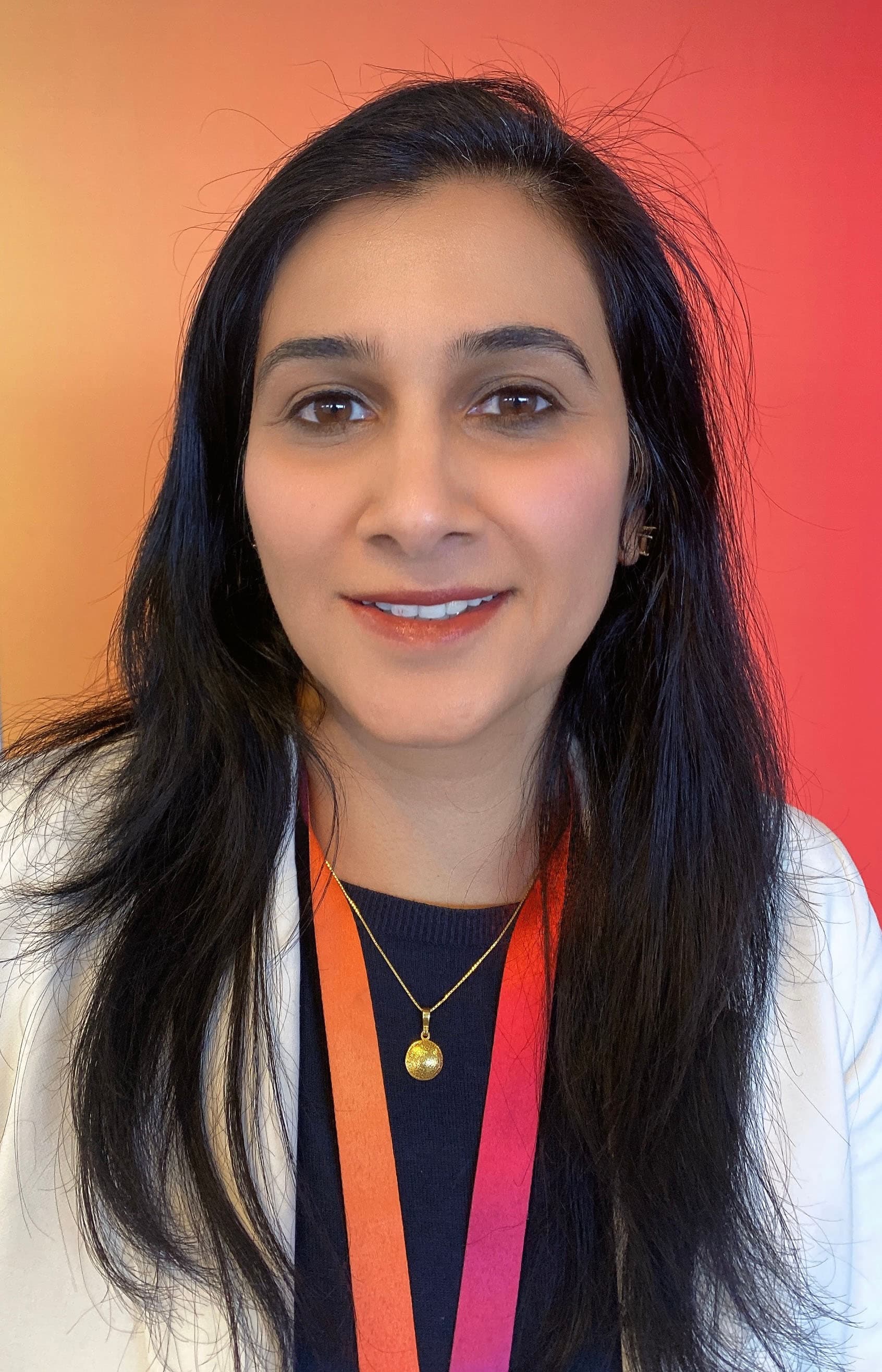 Close-up of a woman smiling, wearing a white blazer and gradient lanyard, in front of a red-orange background.