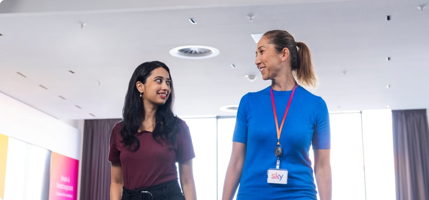 Two women walking indoors and smiling at each other, one wearing a Sky badge and blue shirt, the other in a maroon top.