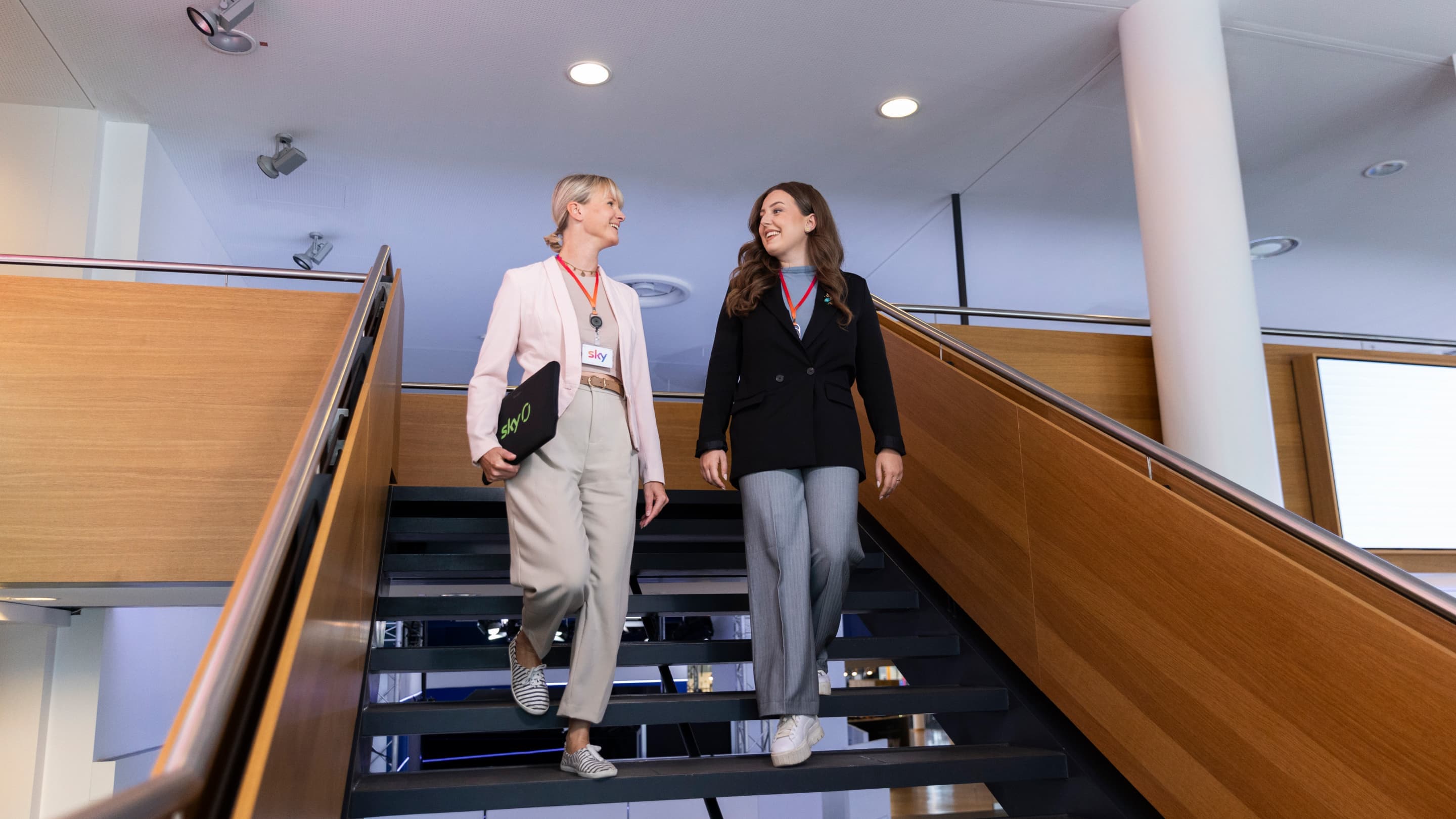 Two women with Sky badges walking down a staircase in a modern office, smiling and holding a Sky-branded laptop sleeve.