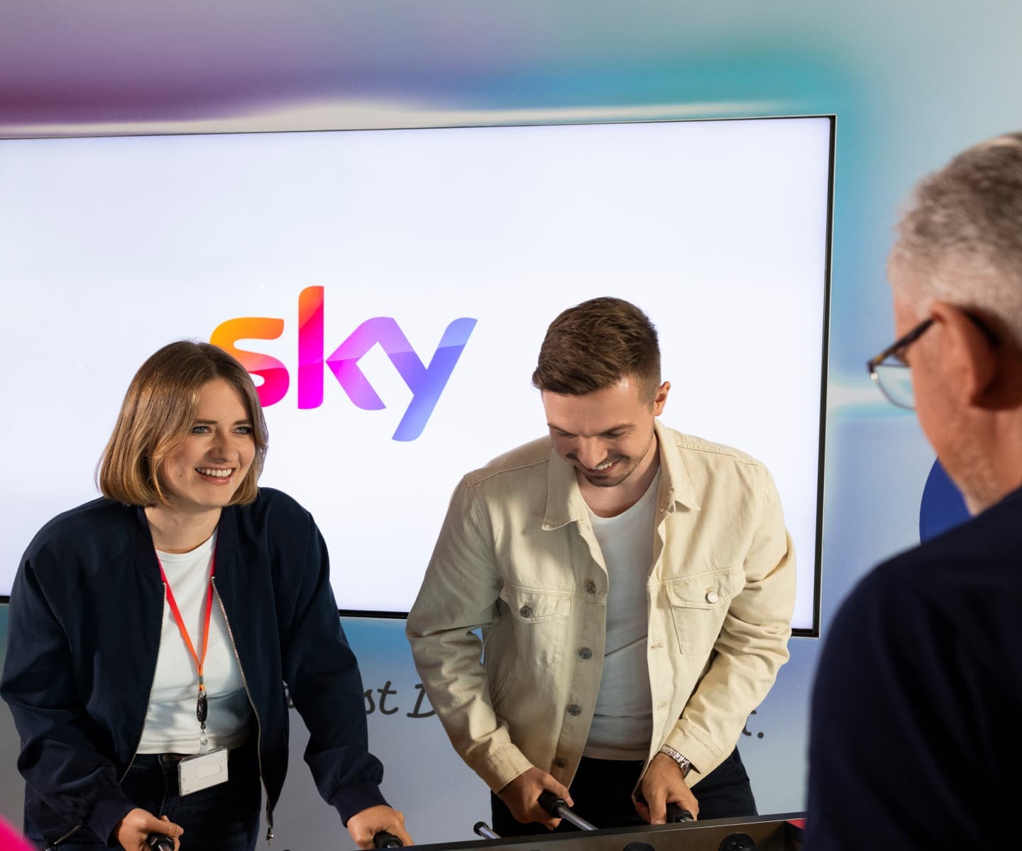 Two colleagues playing table football in front of a screen displaying the Sky logo, with a third person watching nearby.