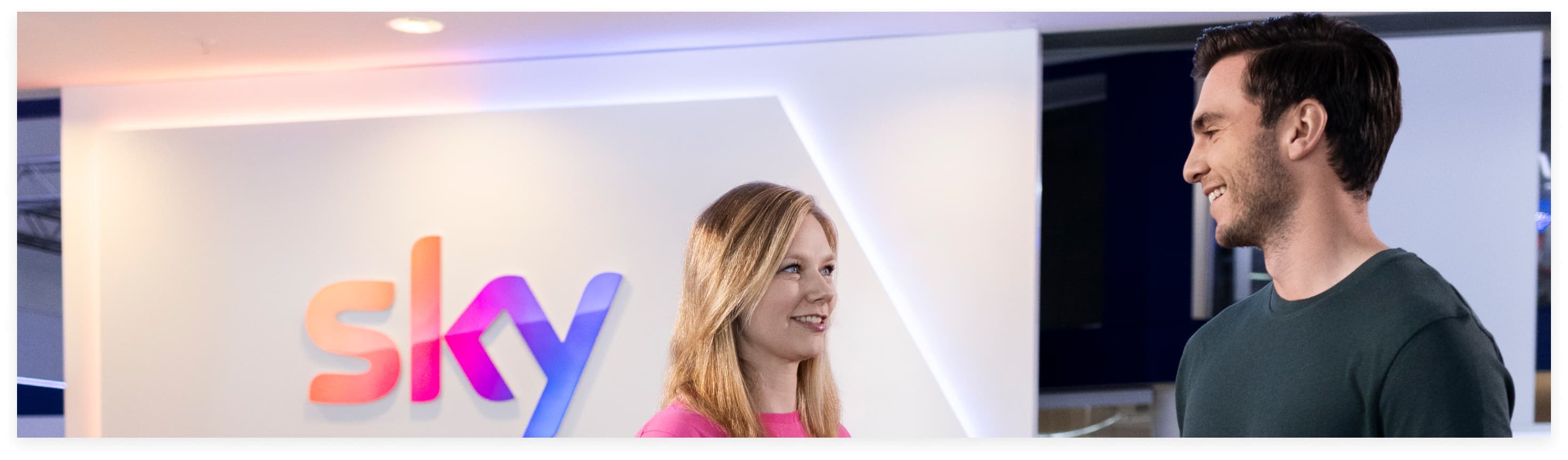 Two colleagues smiling as they talk inside a Sky office, with a large Sky logo on the wall behind them.