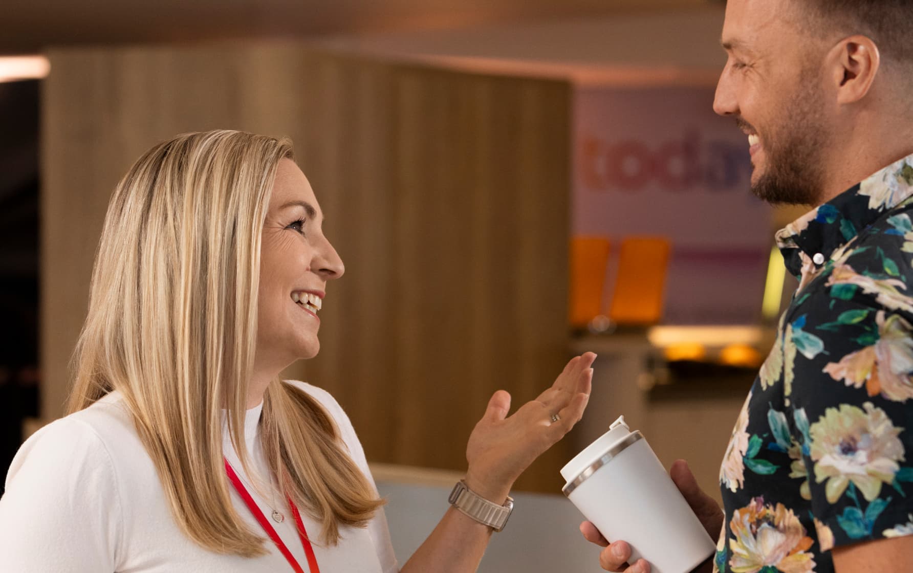 Two colleagues smiling and chatting indoors, one holding a reusable cup, with a blurred office background.