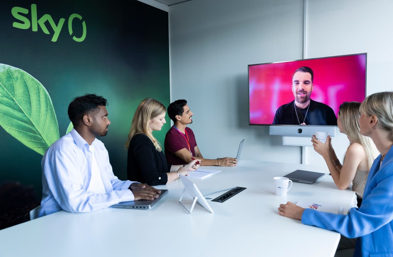 Five people in a modern meeting room with a large “Sky Zero” branding on the wall, attentively watching a colleague speaking via video call on a mounted screen.