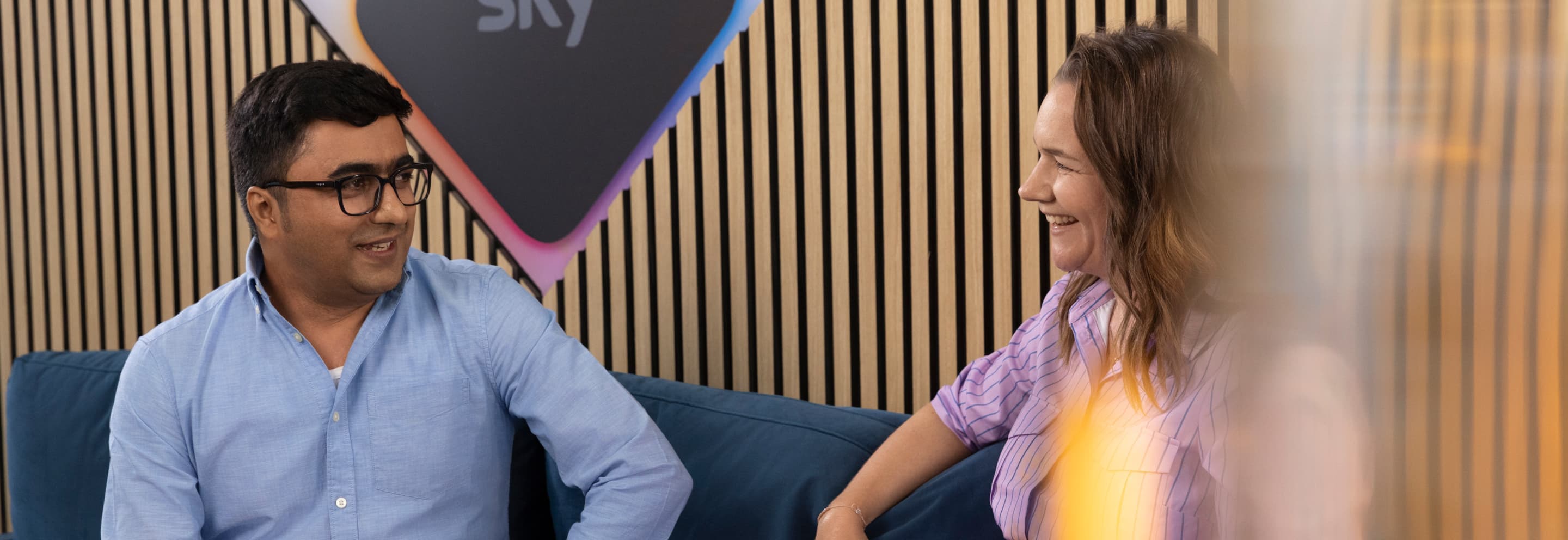 Two colleagues smile and chat on a blue couch in a Sky office with wood panel walls and a Sky logo behind them.
