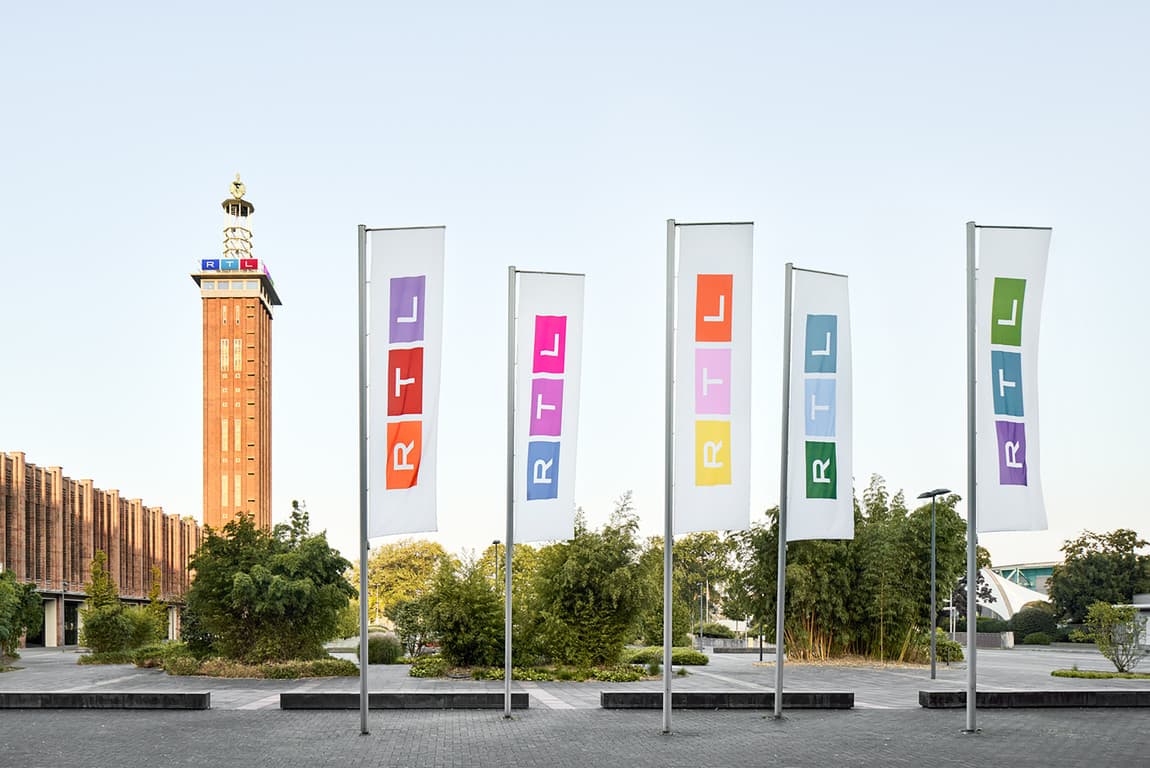 Mehrere Flaggen mit dem RTL-Logo in unterschiedlichen Farben vor einem Gebäude; im Hintergrund ist ein hoher Turm mit einem RTL-Schriftzug zu sehen.